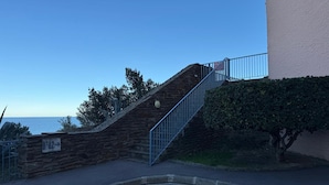 Un escalier en pierre menant à un bâtiment doté d’une rambarde métallique, une enseigne portant l’inscription « B-A-Z », et une vue sur l’océan.