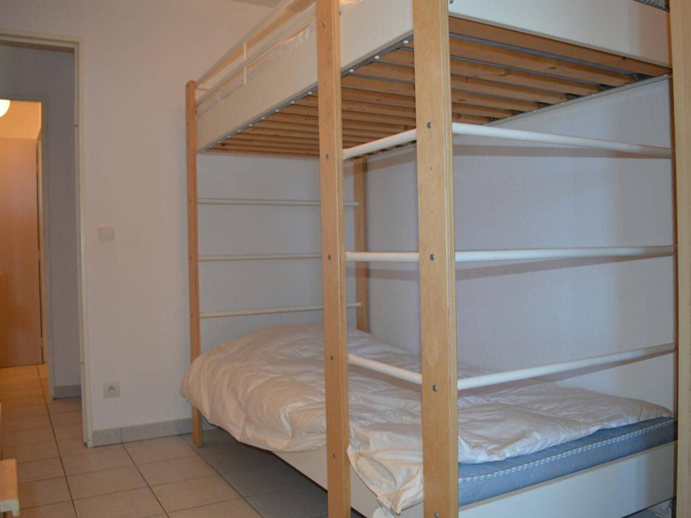 Een slaapkamer met een stapelbed met houten frame, wit beddengoed en een betegelde vloer.