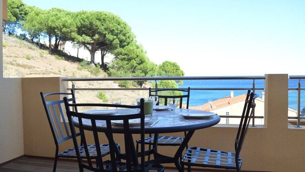 A balcony with a table and chairs, overlooking a coastal area with trees and a view of the sea.