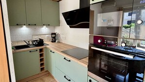 A compact kitchen with green cabinets, a black oven, and a window with curtains. - Cozy 2-Room House with Mezzanine, A/C, Parking, Near Beach - Argeles-sur-Mer (Argelès-sur-Mer)