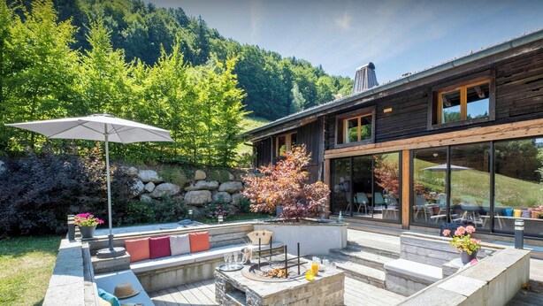 A modern outdoor patio with a fire pit, seating area, and a view of a forested hillside. - Chalet Colombine - luxury base 15p indoor pool & spa, cinema - OVO Network (Thônes)