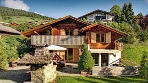A two-story wooden chalet with a balcony, stone foundation, and a well-maintained lawn. - Chalet Le Petit Mazot - romantic break for 4 close to slopes - OVO Network (La Clusaz)