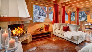 Interior acogedor de una cabaña con chimenea, un sofá, una mesa de comedor y vistas a árboles nevados.