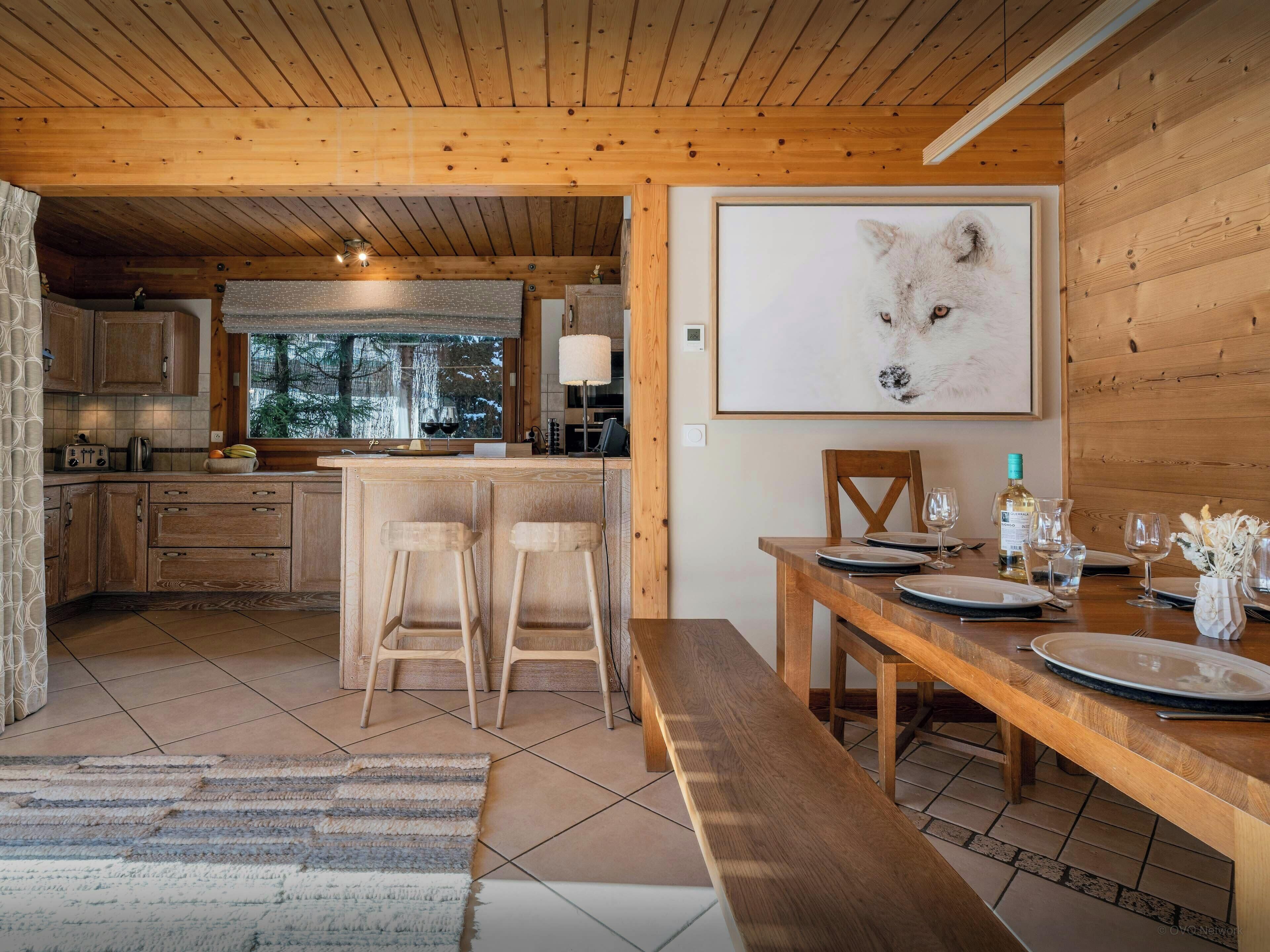 Interior de una cabaña de madera con cocina, comedor y un gran retrato enmarcado de un animal.