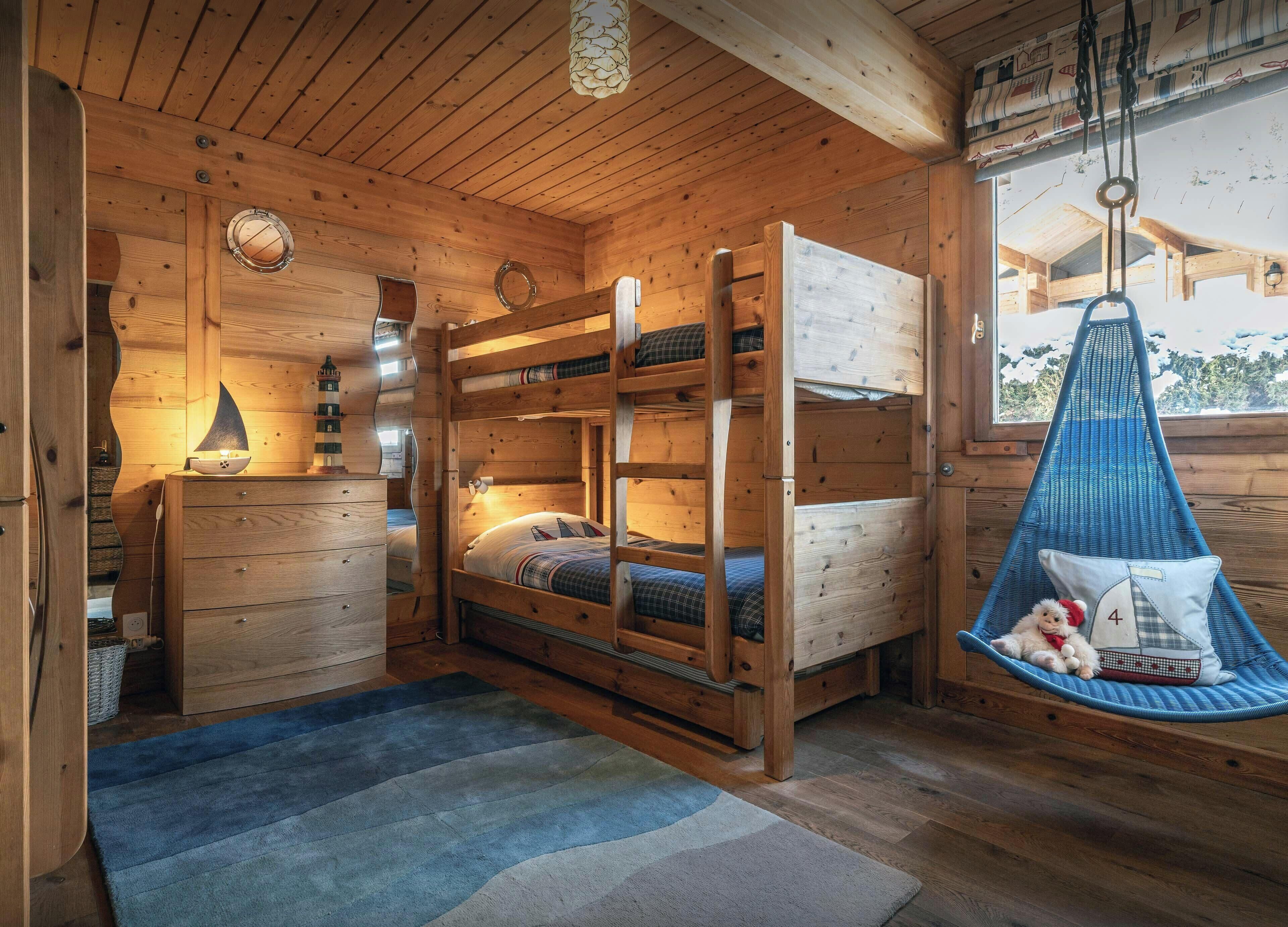 A wooden cabin interior with bunk beds, a desk, a hanging chair, and a window with a view.
