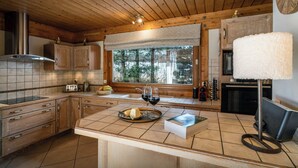 A kitchen with wooden cabinets, a window with a view of trees, a wine glass, and a book on the counter. - Chalet J'Elage - family base 10p, open fire & mountain views - OVO Network (St Jean de Sixt)