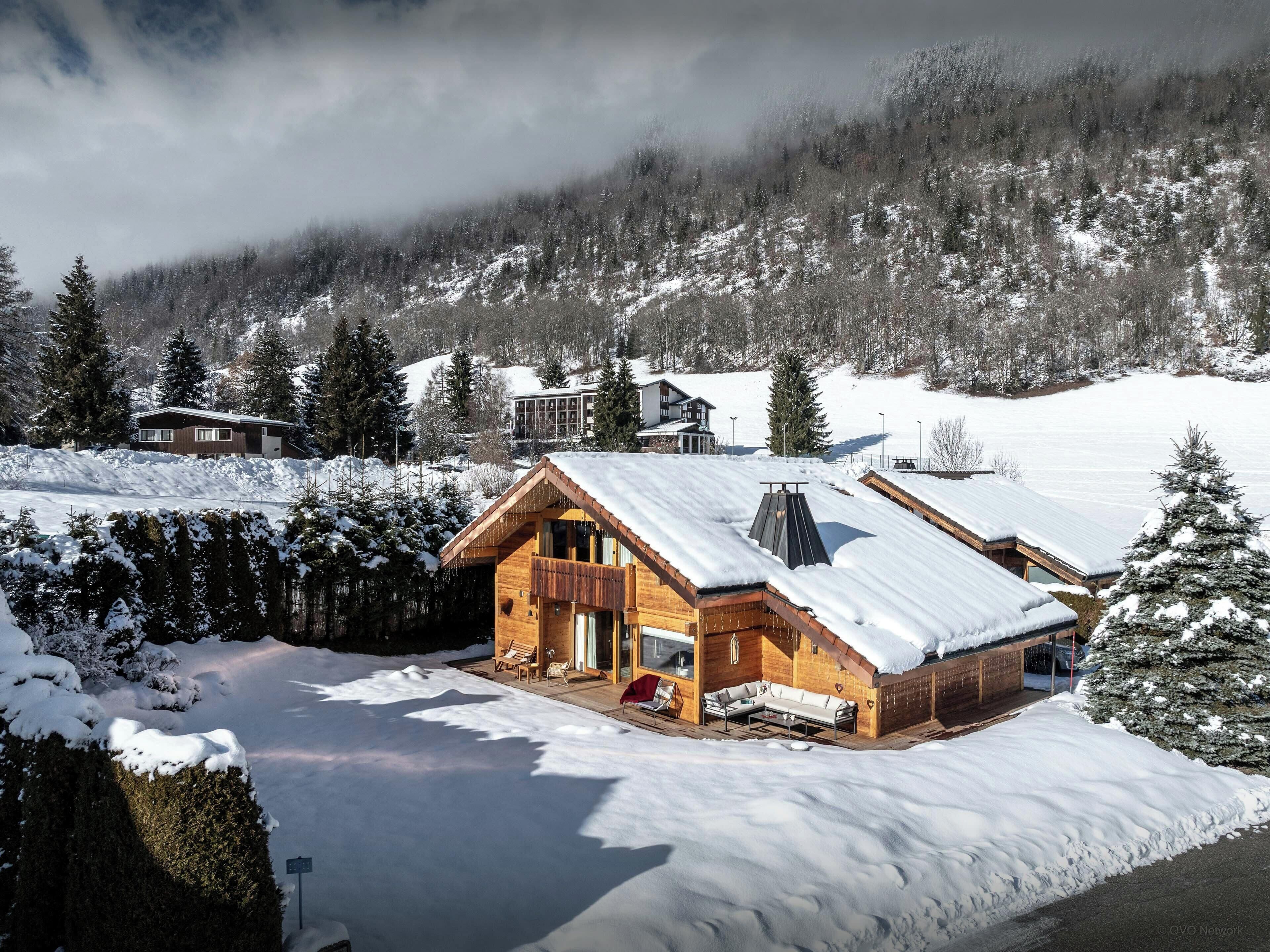A wooden cabin with a sloped roof, surrounded by snow-covered ground and trees.