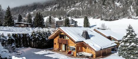 A wooden cabin with a sloped roof, surrounded by snow-covered ground and trees.