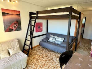 A bunk bed room with a ladder, a sofa, and a chair. There is a painting on the wall and a patterned floor.