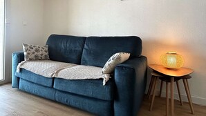 En stue med en mørkeblå sofa, puder med mønster, et lille bord med en lampe og et billede på væggen.