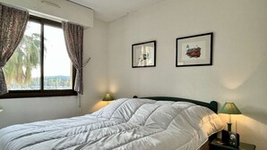 Una cama bien hecha con ropa blanca, un cabecero verde y dos lámparas de noche. Hay dos cuadros en la pared y una ventana con cortinas.