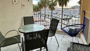 Un balcón con una mesa y sillas de vidrio, una silla azul, y vista a un puerto deportivo con barcos y palmeras.