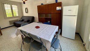 A small room with a table, chairs, a sofa, a refrigerator, and a window with blinds.