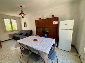 A small room with a table, chairs, a sofa, a refrigerator, and a window with blinds. - Cozy 2-Bedroom House, 150m from Beach, Terrace, and Parking (Port-la-Nouvelle)