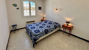 A bedroom with a bed, two bedside tables, a small stool, a window with bars, and a wall-mounted light fixture.