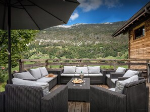 Une terrasse en bois, meublée de meubles en osier, avec une table dressée où l’on a disposé du pain et des verres à vin.