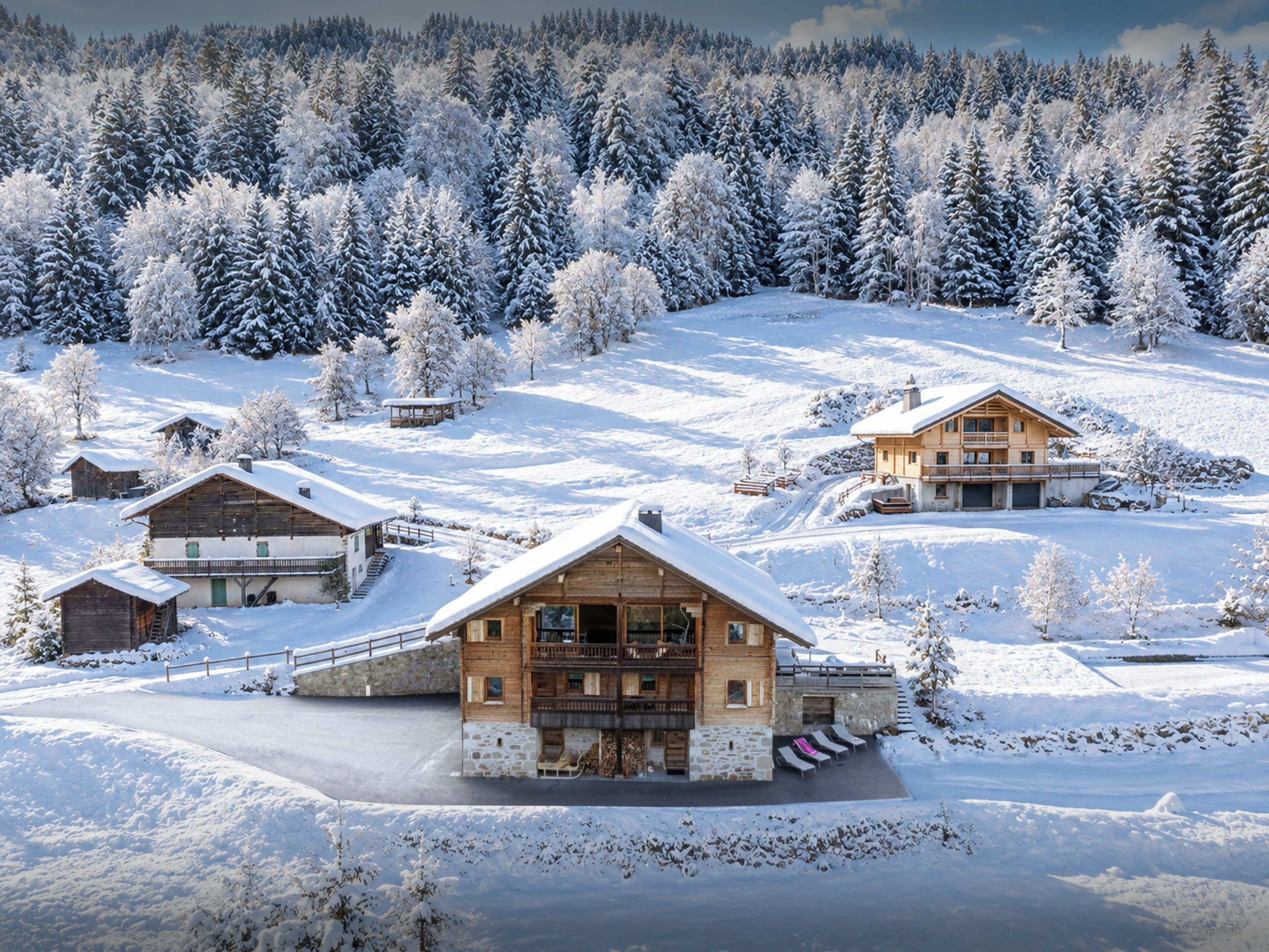 Un chalet de montagne enneigé, composé de plusieurs bâtiments, avec une piste de ski et un paysage boisé en arrière-plan.
