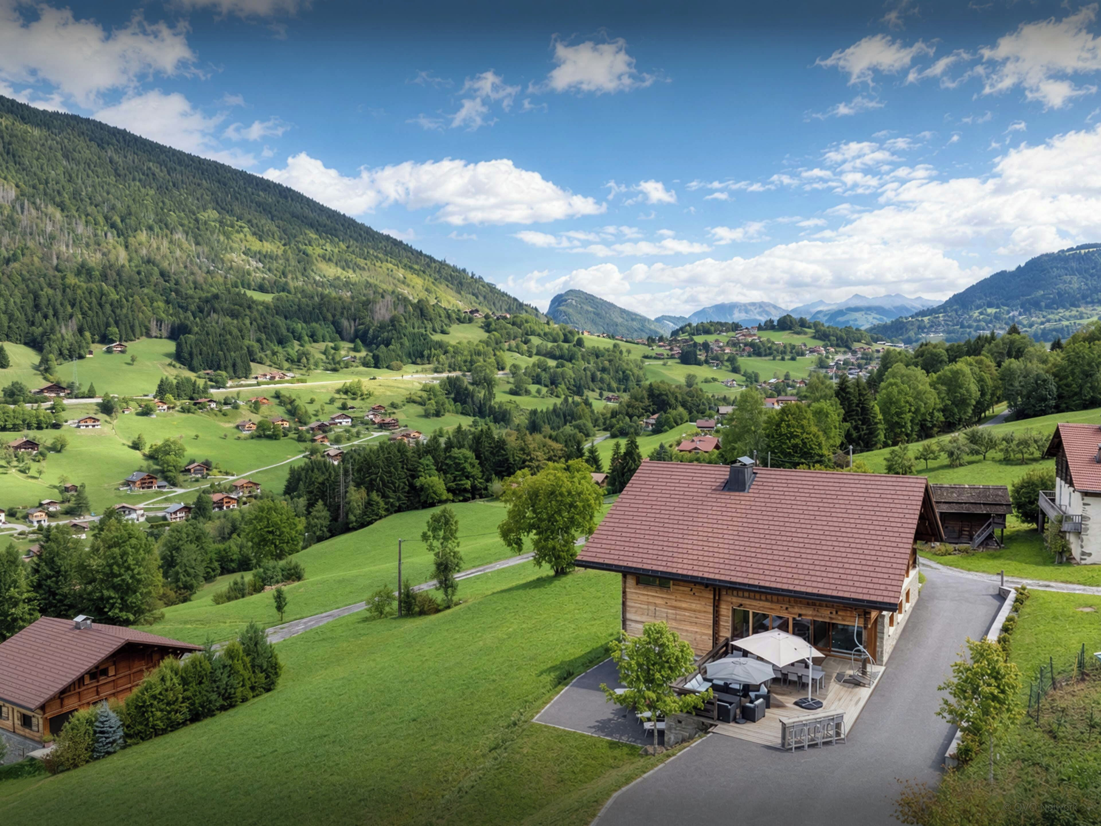 A mountain lodge with a patio area, surrounded by lush green fields and forested hills.