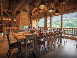 Un intérieur en bois avec une table à manger dressée pour quatre personnes, entourée de chaises, et de grandes fenêtres offrant une vue sur un paysage boisé.