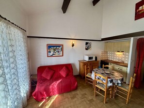 A compact living space with a red sofa, a dining table with chairs, a kitchenette, and a painting on the wall.