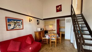 A cozy living room with a red sofa, a wooden dining table with chairs, and a kitchen area with a microwave. - House 2 rooms, cabin, 6 pers, 400m from beach, parking, terrace (Port-la-Nouvelle)