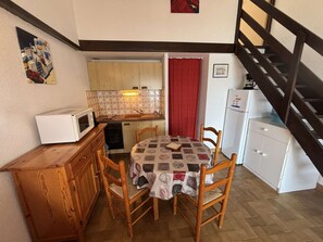 A compact kitchen with a microwave, sink, and refrigerator. There is a dining table with chairs and a red curtain. A wooden cabinet and a staircase are also visible.