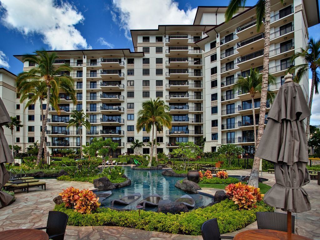 Un hotel con varias plantas, piscina al aire libre, palmeras y sillones de descanso.