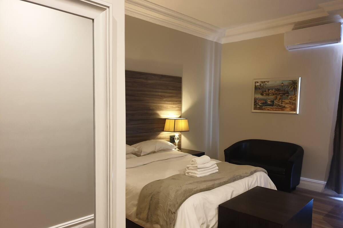 Een hotelkamer met een bed, nachtkastje, bureau, stoel, lamp en een schilderij aan de muur.
