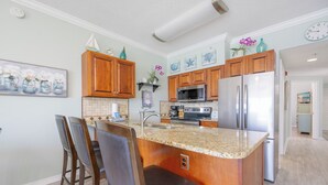 A kitchen with wooden cabinets, a granite countertop, a stainless steel refrigerator, and a bar area with chairs. - Royal Palms 144 Beachfront Gulf Escape  Pool, Hot Tub & Sauna (Gulf Shores)