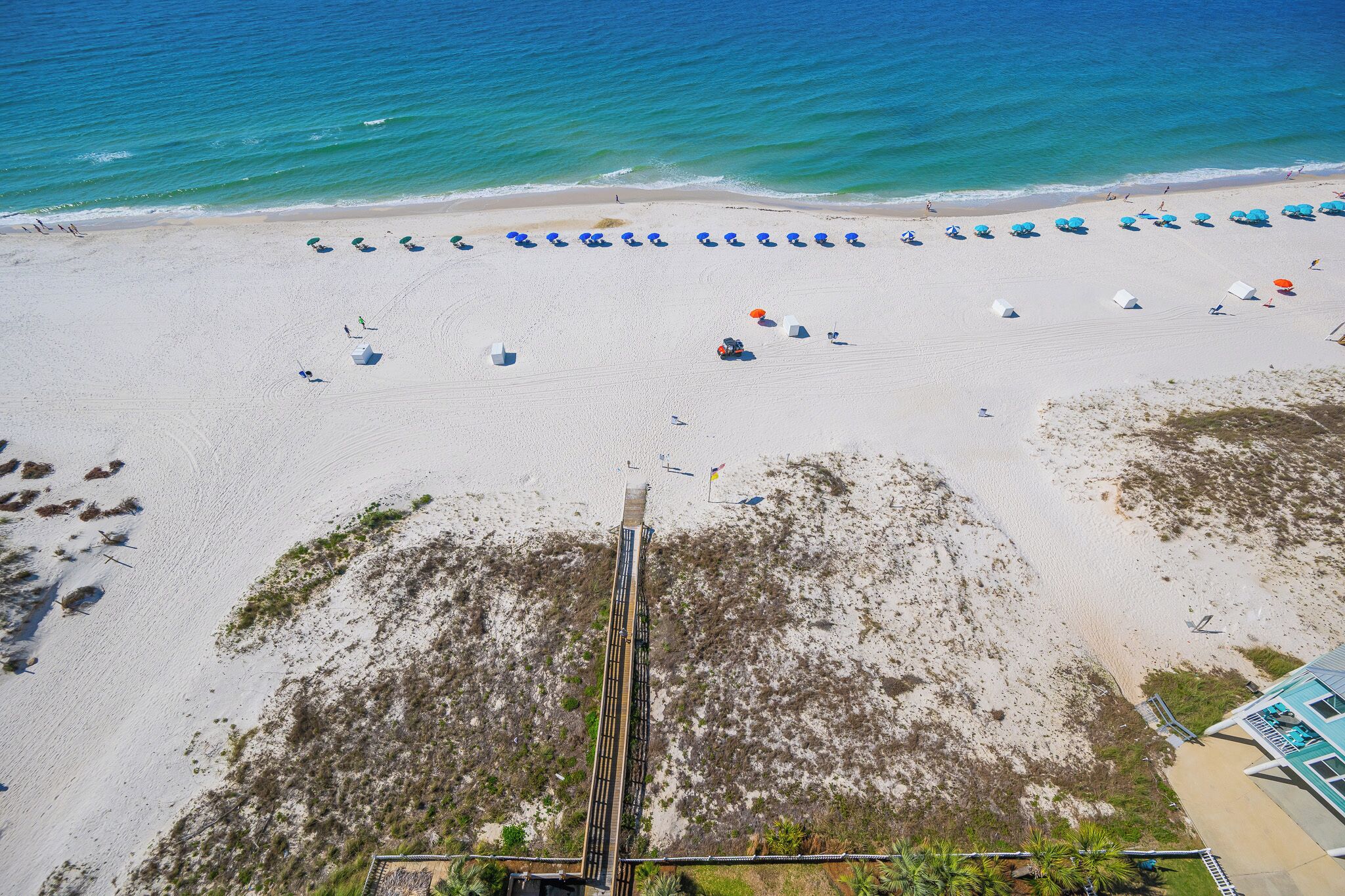 Vista aérea de una playa con hileras de sombrillas y sillas de playa, arena y una estructura con una escalera que baja.