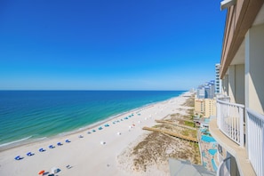 A beach view from a balcony with a clear sky, a sandy beach with umbrellas, and a pool area.