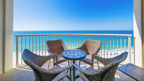 A balcony with wicker chairs and a table, overlooking a beach with clear blue water and a white railing. - Royal Palms 144 Beachfront Gulf Escape  Pool, Hot Tub & Sauna (Gulf Shores)