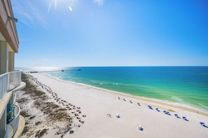 A beach with a pier extending into the ocean, a sandy beach with umbrellas, and a building with a balcony overlooking the scene. - Royal Palms 144 Beachfront Gulf Escape  Pool, Hot Tub & Sauna (Gulf Shores)
