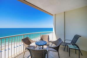 A balcony with wicker furniture and a table, overlooking a beach with a clear view of the ocean.