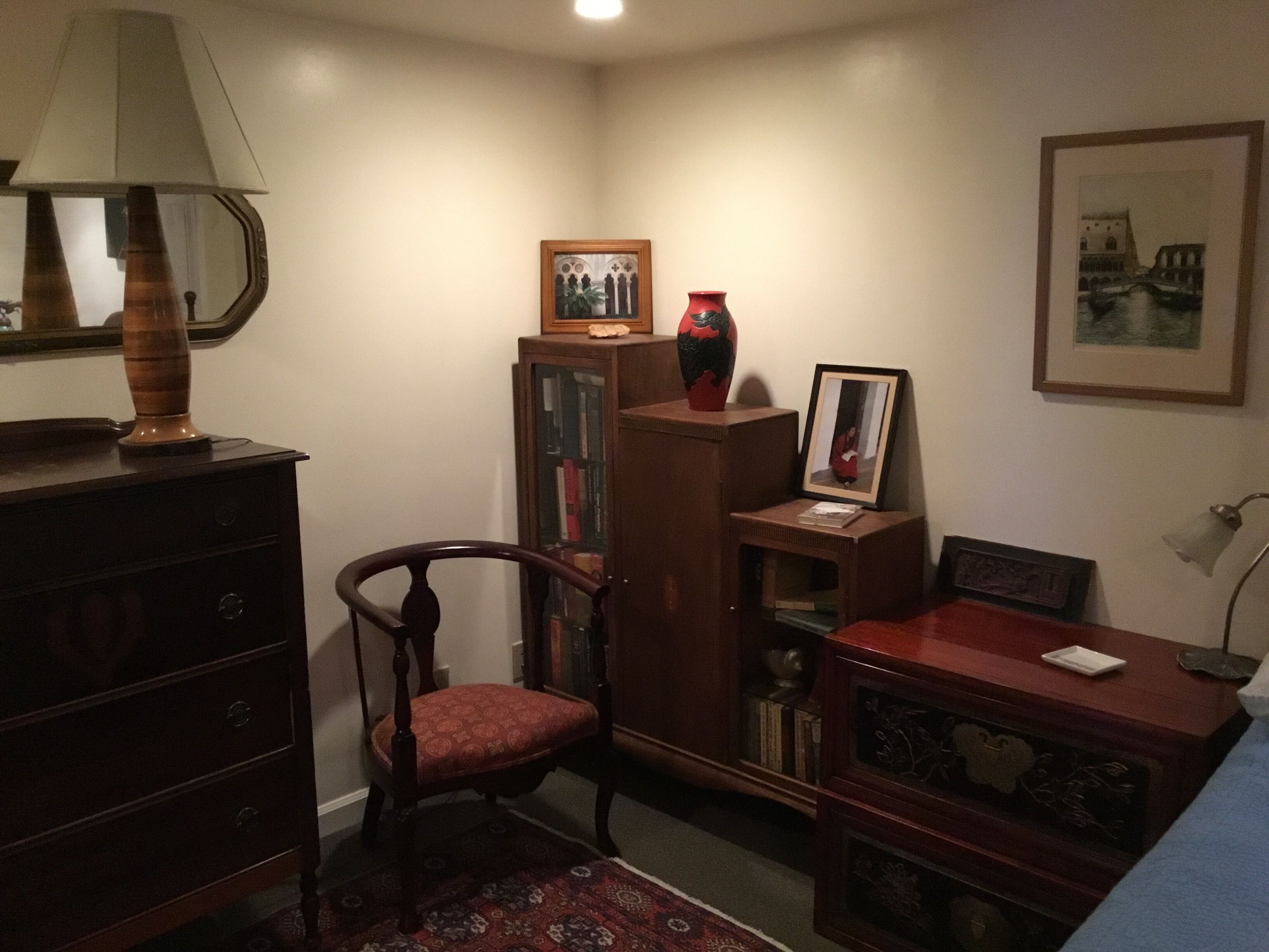 Une pièce d’angle avec une chaise en bois, une bibliothèque remplie de livres, une lampe et un tableau encadré accroché au mur.
