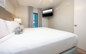 A hotel room with a large bed, a flat-screen TV mounted on the wall, and a view of the outside through a window with curtains.