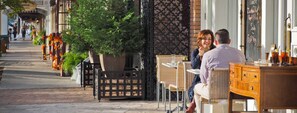 A couple dining at an outdoor cafe with wicker chairs and a wooden table, a black metal fence, and a brick wall in the background. - Best Location in Town -  Steps from downtown, wine shuttles, restaurants, etc. (Fredericksburg)