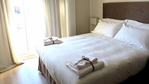 A neatly made bed with white linens and towels, a bedside lamp, and a view of the outdoors through a window with curtains. - Gatto Bianco Le Terrazze (Bari)