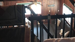 A room with a sloped wooden ceiling, a ceiling fan, a window, and a staircase leading to an upper level.