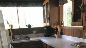 A compact kitchen with wooden cabinets, a white countertop, a sink, and a window with a view of greenery.