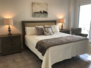 A bedroom with a large bed, two bedside tables with lamps, a chair, and a painting on the wall.