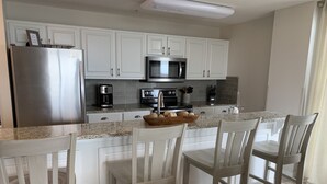 A modern kitchen with white cabinets, a stainless steel refrigerator, a microwave, and a coffee maker. There are four wooden chairs around a table.