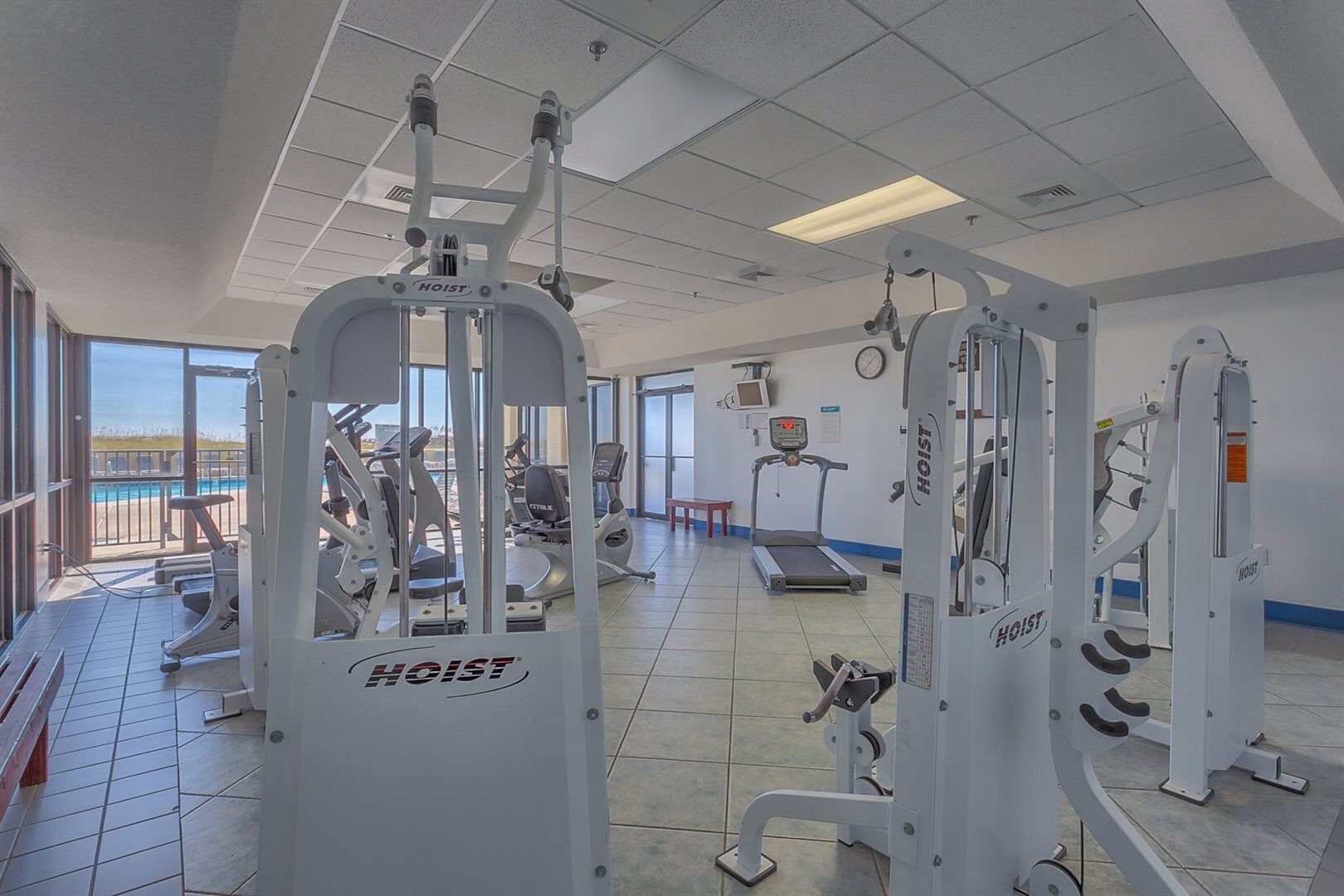 Une salle de sport équipée de divers appareils d’exercice, notamment un appareil à câbles et un tapis de course, et offrant une vue sur une piscine à l’extérieur.