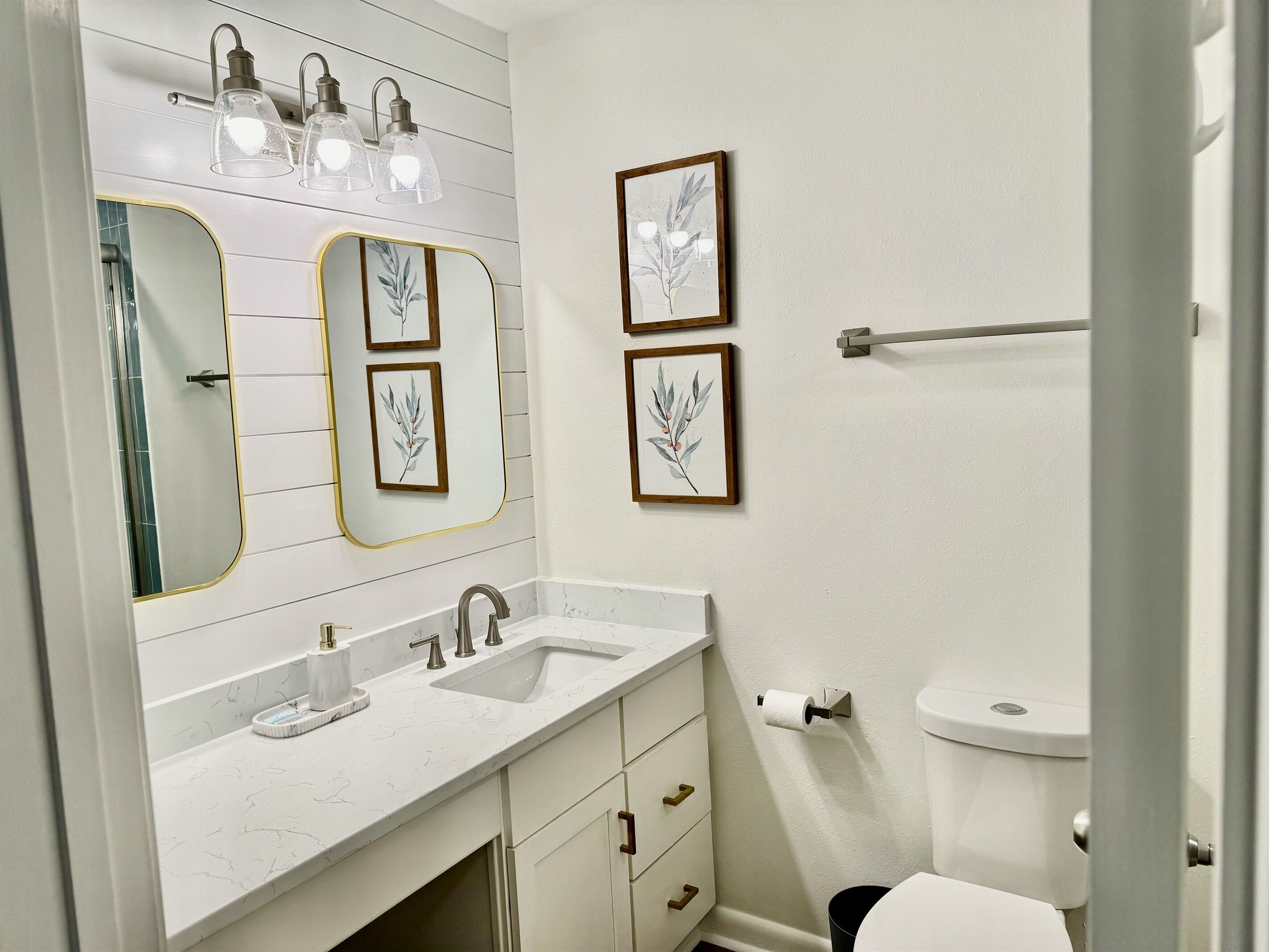 A bathroom with a white vanity, two sinks, a large mirror with gold trim, and framed botanical prints on the wall.