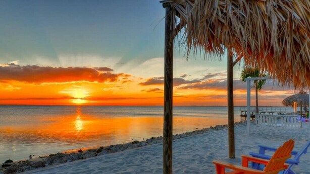 Eine Strandansicht mit Sonnenuntergang, Holzbauten und Liegestühlen.