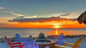 Sonnenuntergang über einem Sandstrand mit einem Lagerfeuer und bunten Adirondack-Stühlen.
