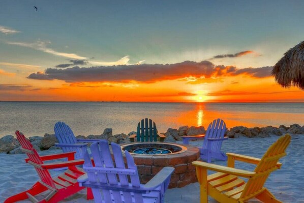A sunset view with a fire pit and colorful Adirondack chairs on a sandy beach.
