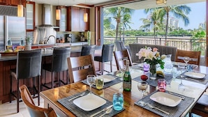 A dining area with a wooden table set for four, surrounded by chairs. A kitchen with modern appliances and a large window overlooking palm trees is visible in the background. - Fall Specials & Sales now-BT307-Beautiful pool and ocean views (Kapolei)