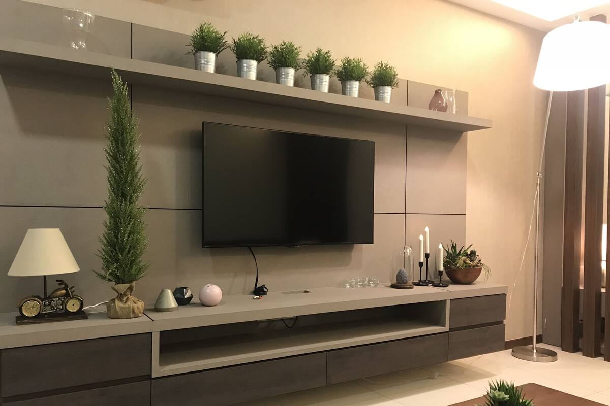 Un salon moderne avec un téléviseur à écran plat fixé à un mur gris, agrémenté d’objets décoratifs tels que des plantes en pot, des bougies et une petite horloge.