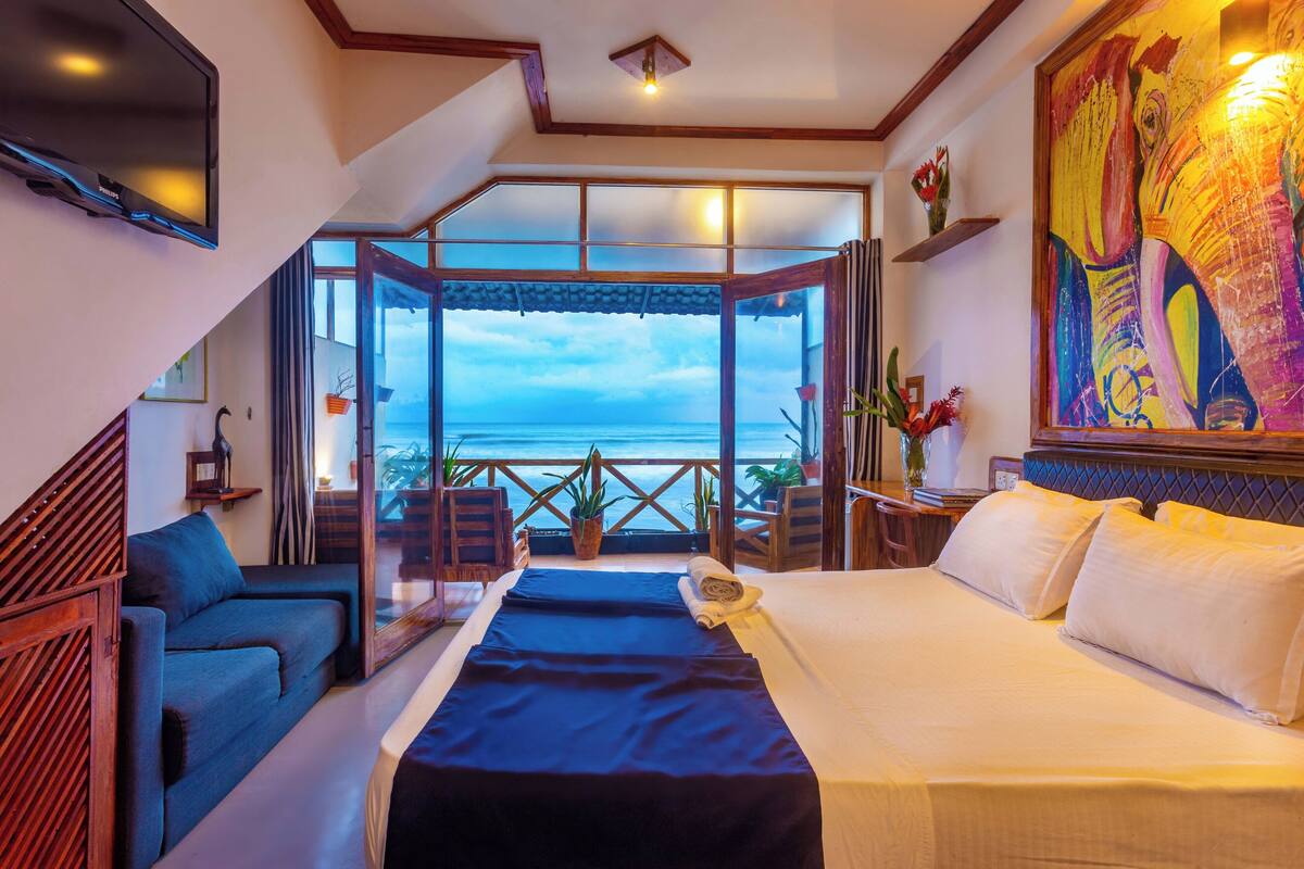 A hotel room with a bed, a sofa, a TV, a painting, and a balcony with a view of the sea.